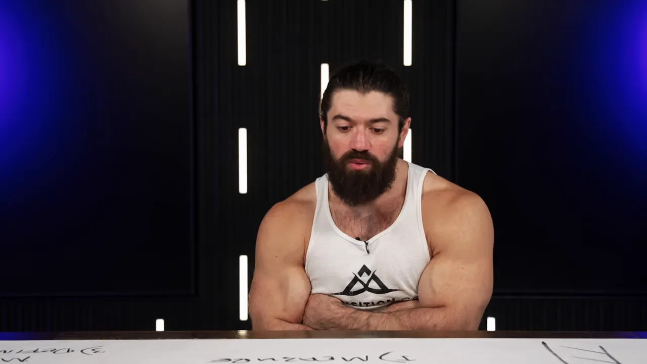 Presenter looking down at the whiteboard surface with a focused expression, seated at a desk with studio lights and vertical accents behind.
