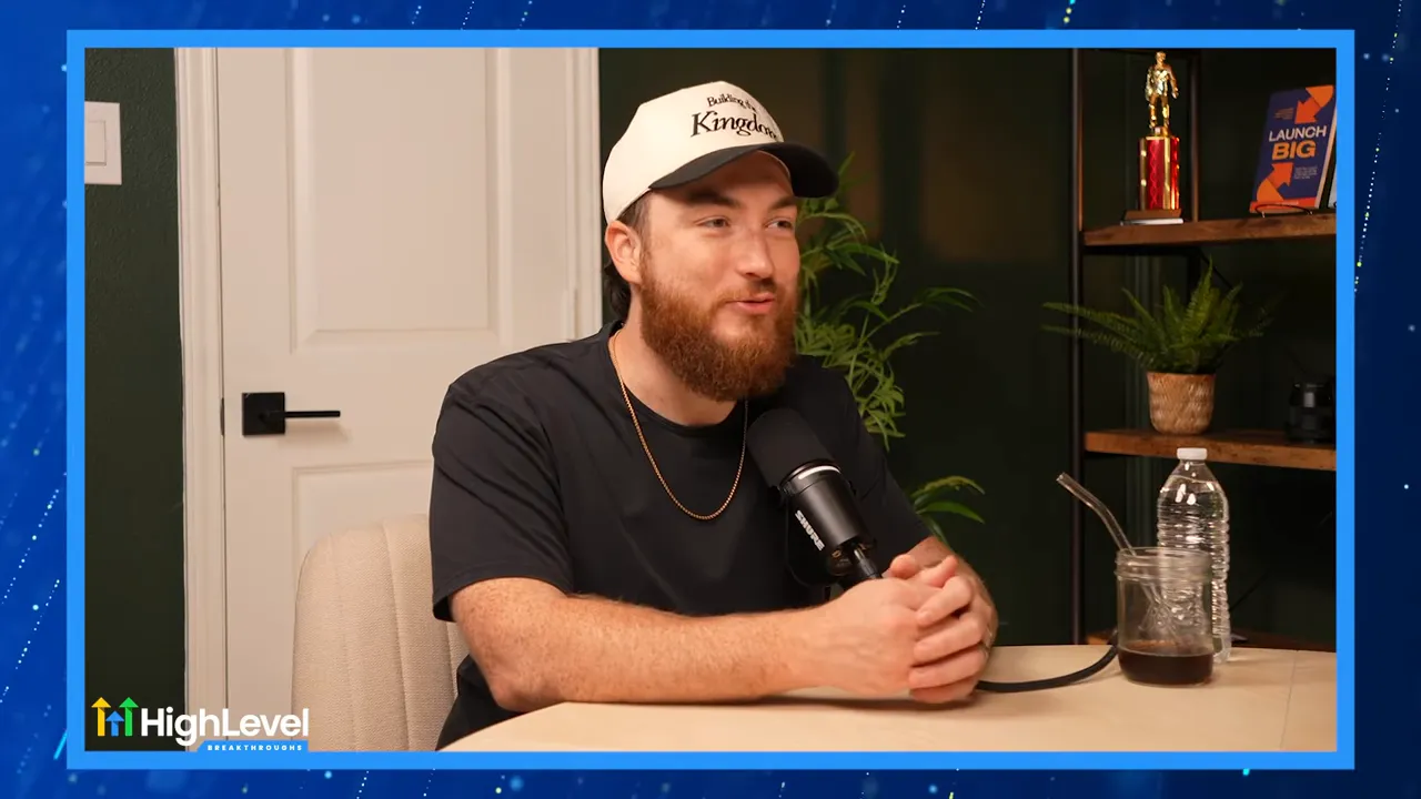 Podcast guest leaning forward and speaking into a microphone with hands clasped on the table; clear, well-framed shot with background shelf and plant.