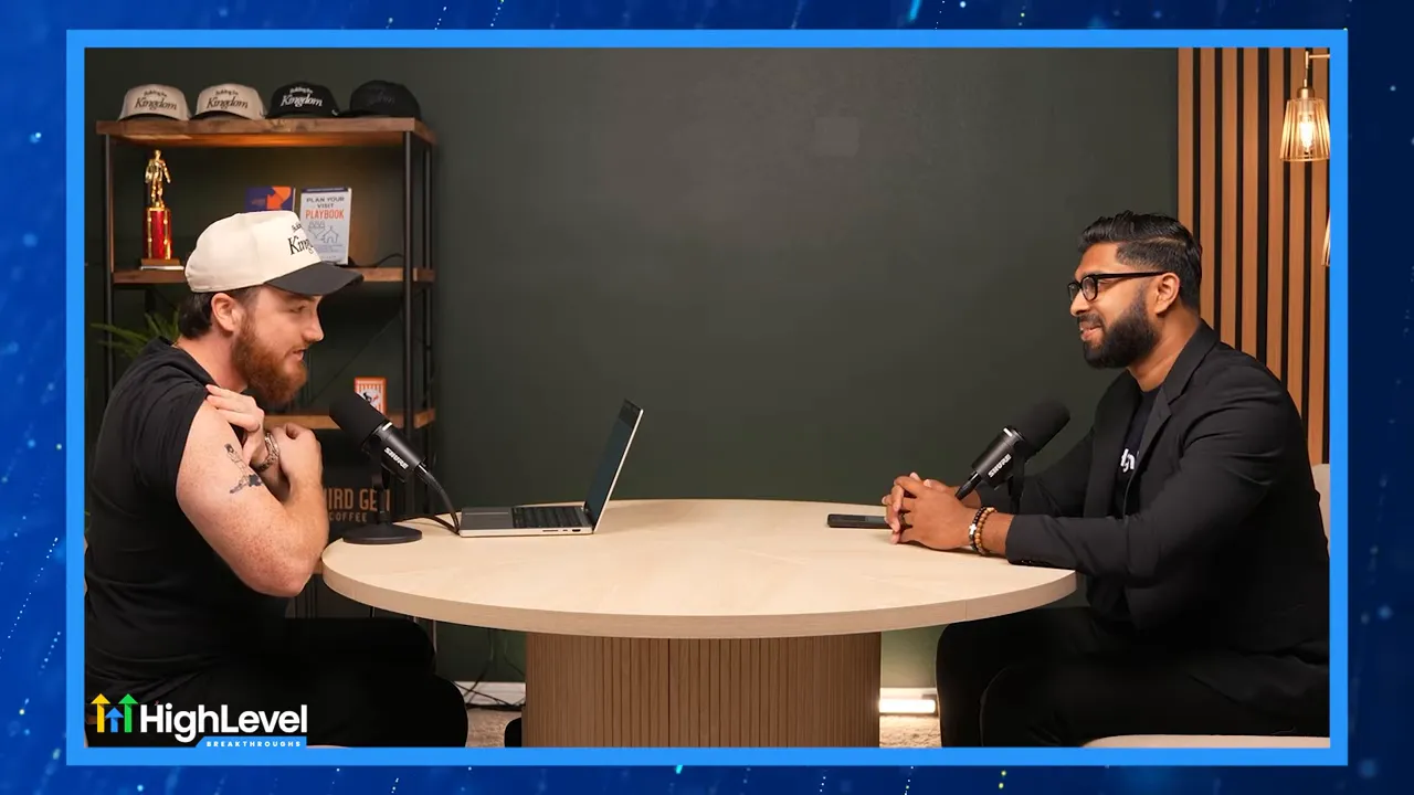 Clear two-shot of two podcast hosts seated at a round table with microphones and a laptop, symmetrical composition and neutral background.