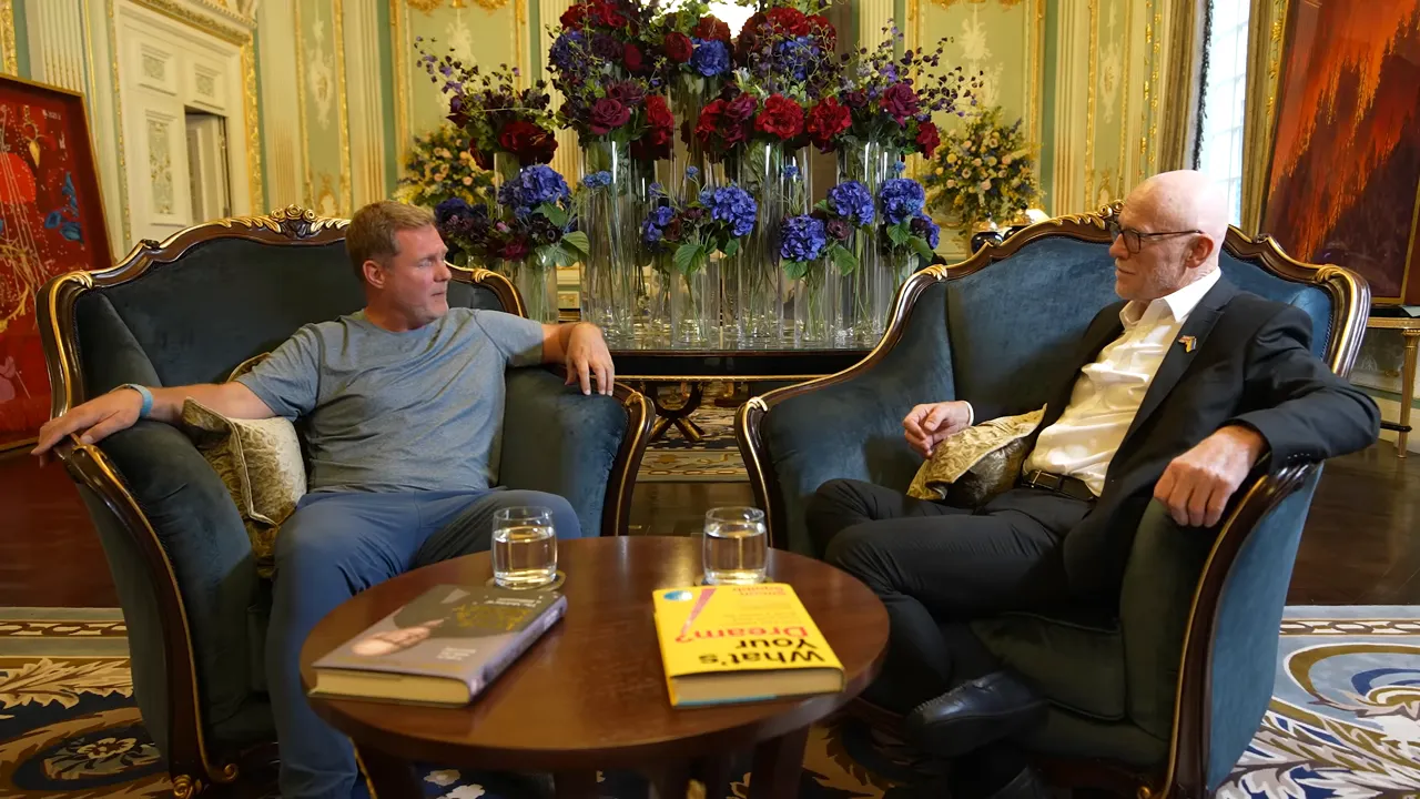 Two men seated in armchairs facing each other across a small table with books and water glasses, floral display behind them.