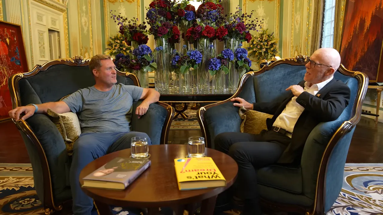 Two people seated across from each other in armchairs with books and glasses on a table and a large floral arrangement behind them, in an ornate interior.