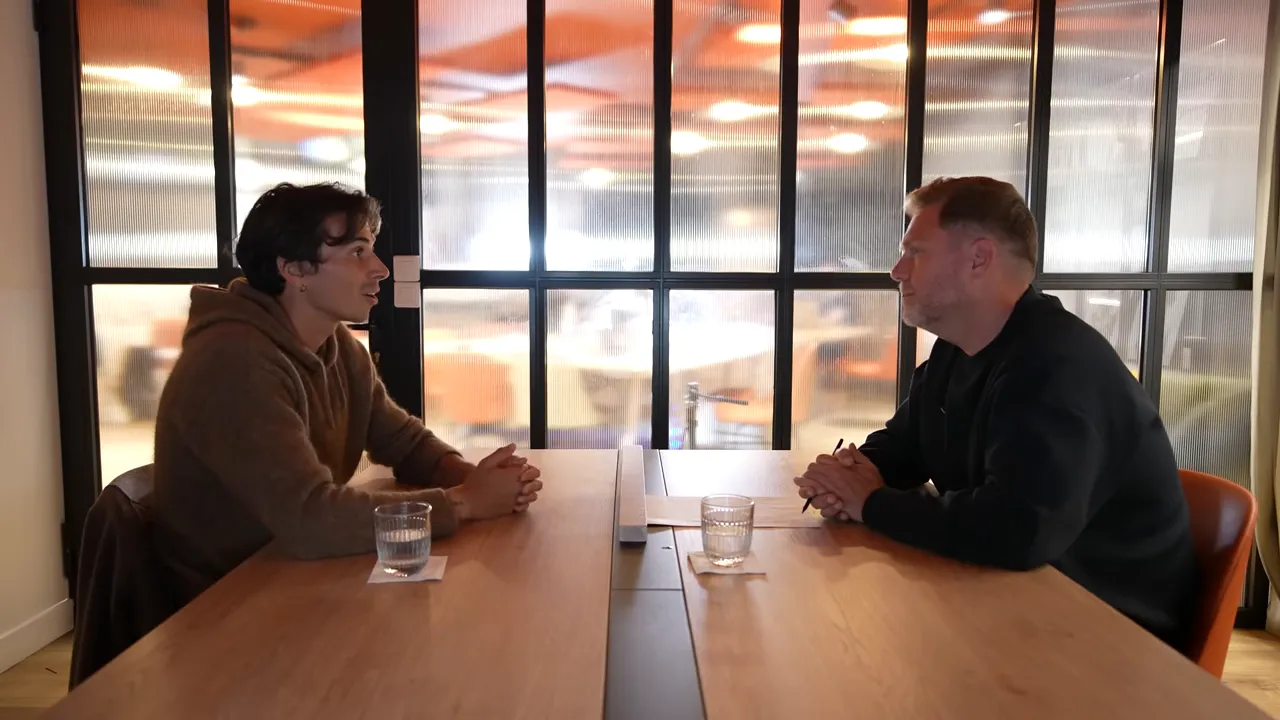 Two people sitting across a wooden table in a meeting room, discussing while facing each other with a glass partition behind them.