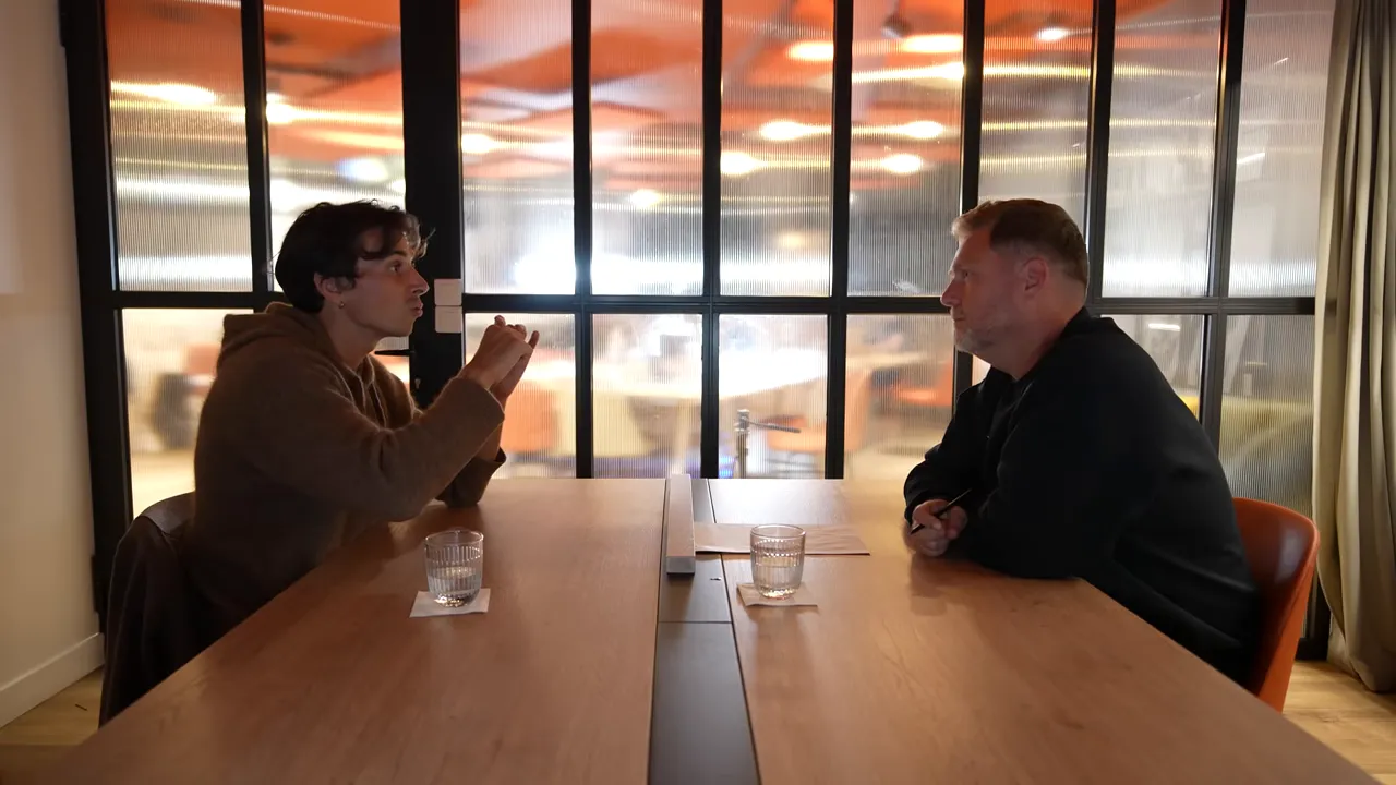 Two people facing each other across a wooden table in a meeting room with papers and glasses on the table, mid-conversation.