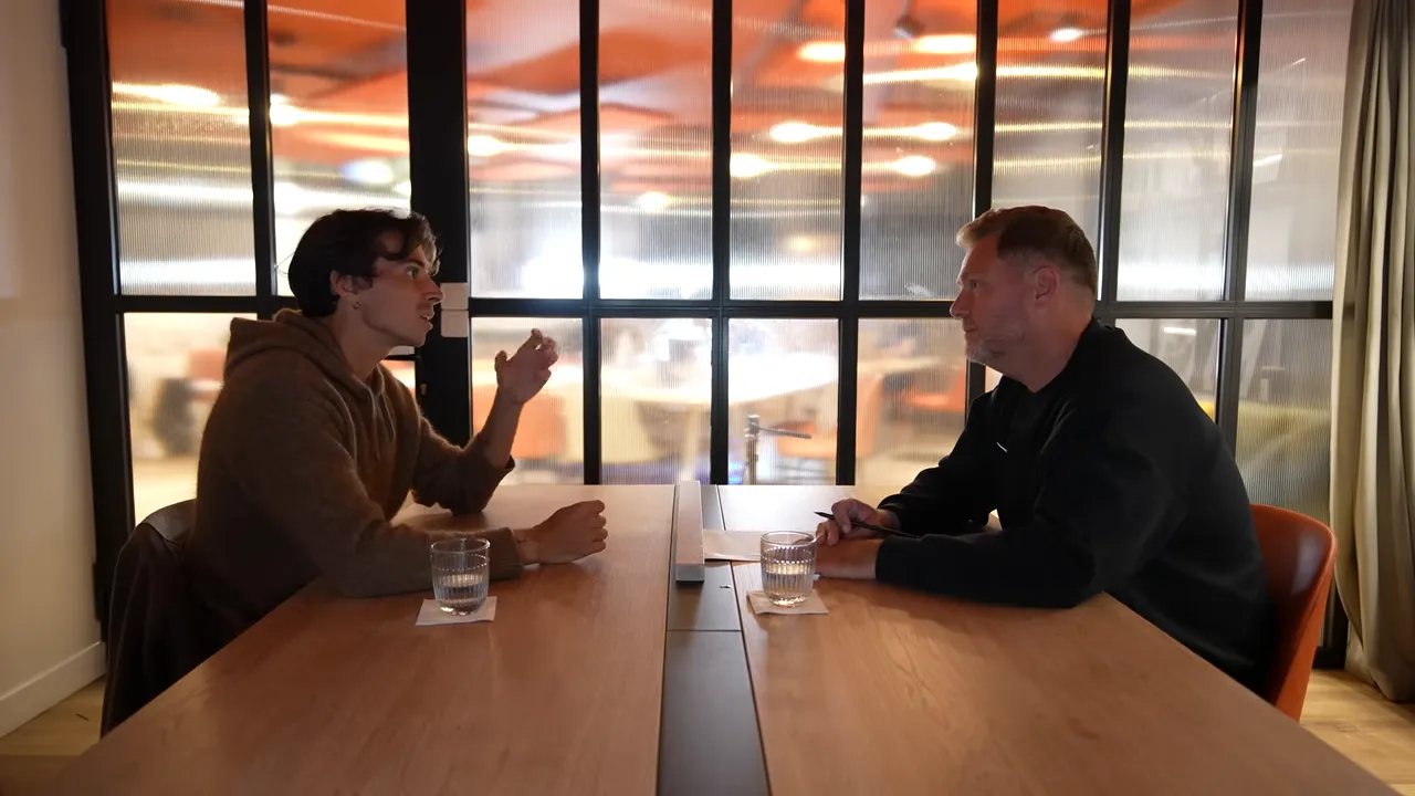 Wide shot of two people facing each other across a table in a meeting room with a glass partition behind them, one person gesturing while speaking.