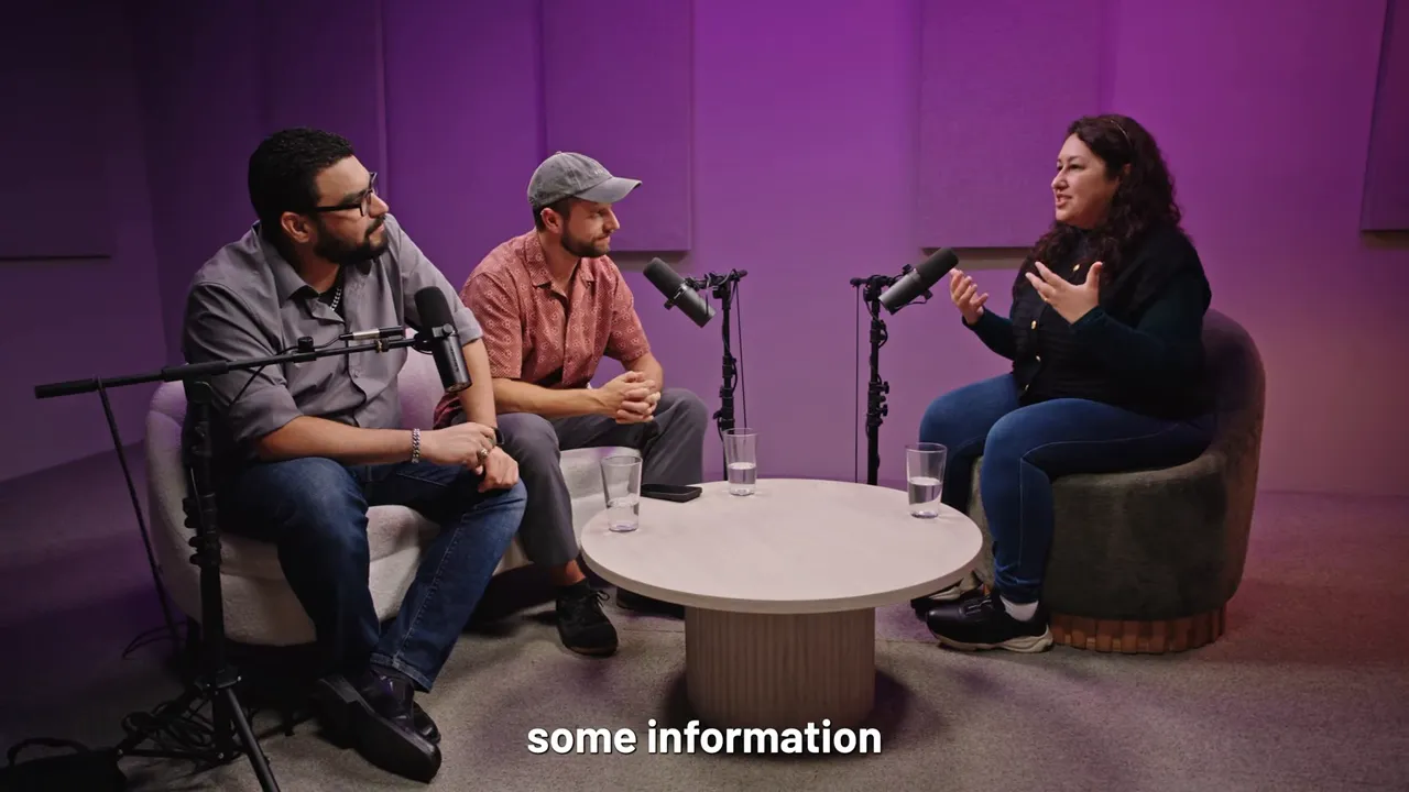 Wide studio shot of three hosts in conversation around a table and microphones