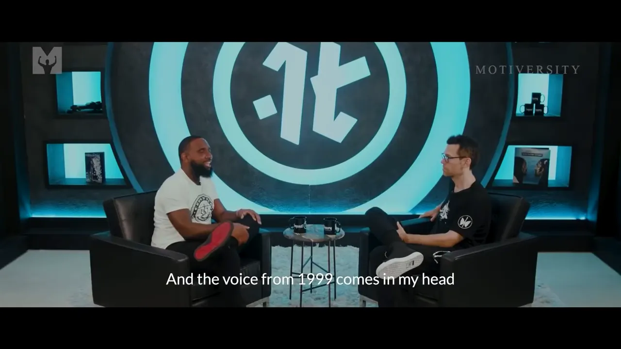 Wide shot of two-person studio interview with circular logo backdrop; guest on left and host on right, subtitle reads 'And the voice from 1999 comes in my head'.