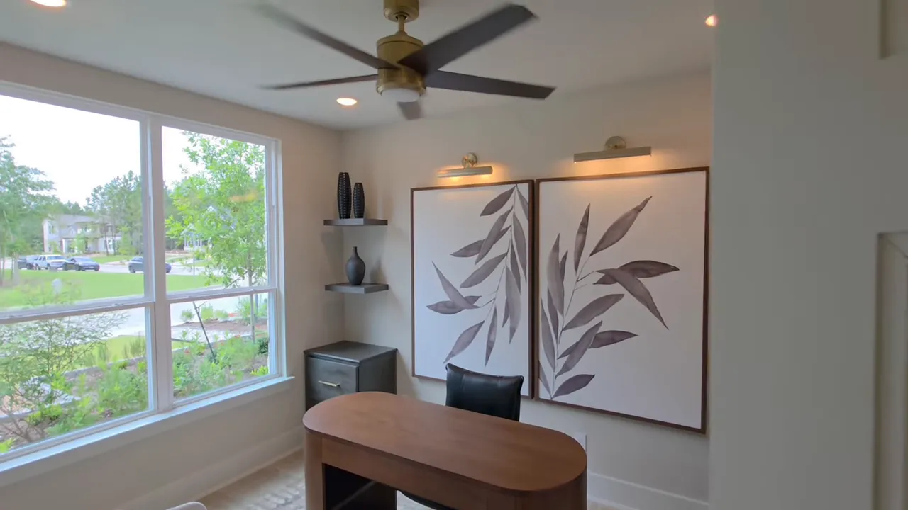 Small home office with wooden desk, large window, botanical wall art and ceiling fan