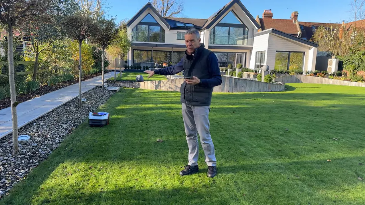 Reviewer pointing to Navimow i105 navigating near a raised garden bed and pebble border