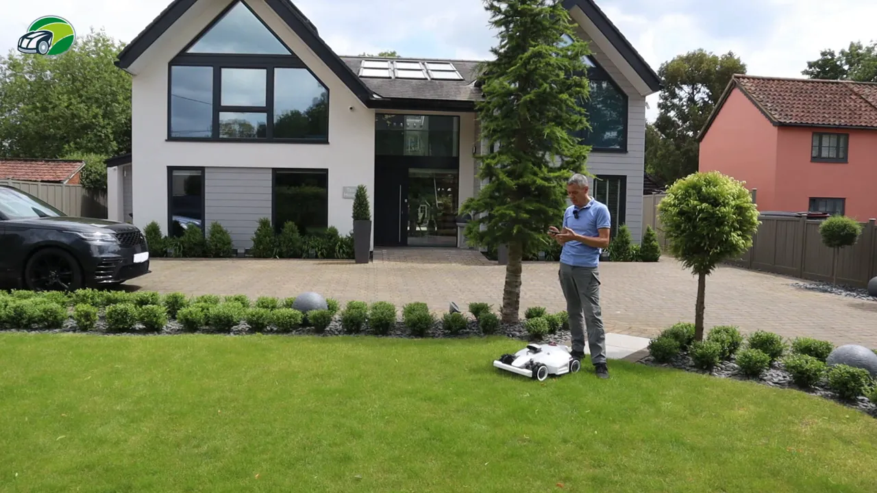 Luba 2 mower on lawn beside a paved driveway while reviewer checks the app to map a channel