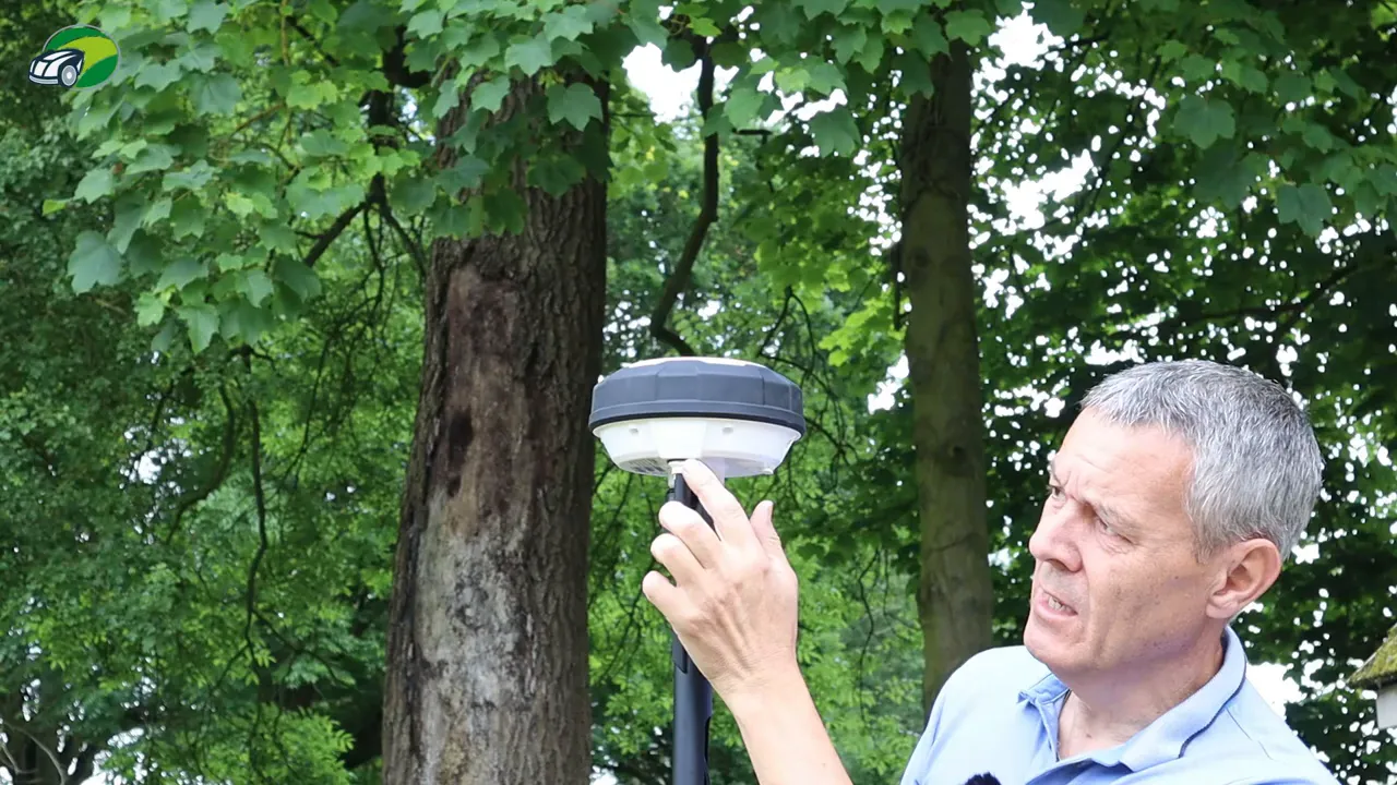 Reviewer demonstrating the RTK station top cover by hand on a pole-mounted GNSS antenna with trees in the background.