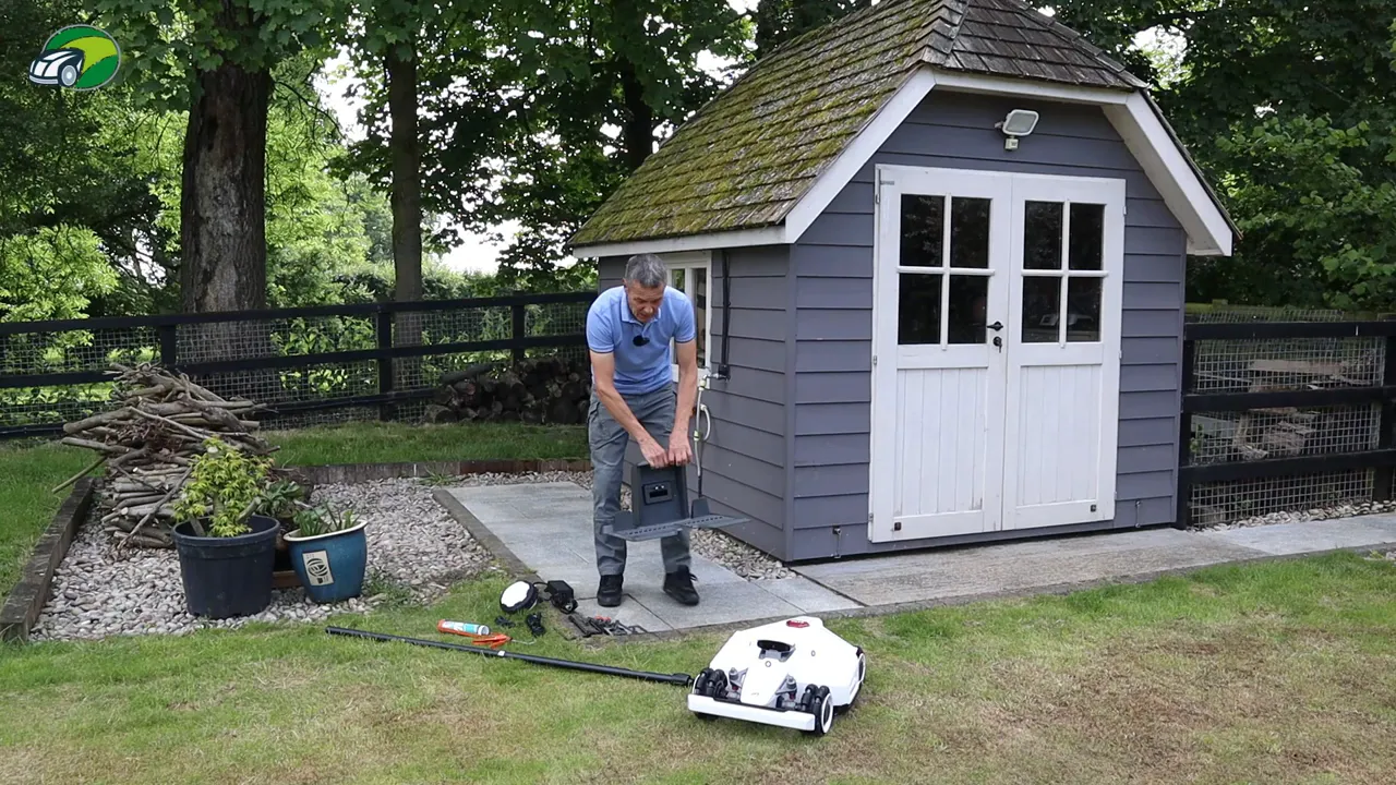 Technician lifting and positioning the RTK charging base beside a garden shed with the Luba 2 mower visible in the foreground.