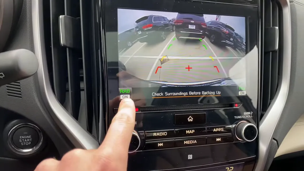 Infotainment backup camera screen with a finger pressing the RAB ON button and the message 'Check Surroundings Before Backing Up'.