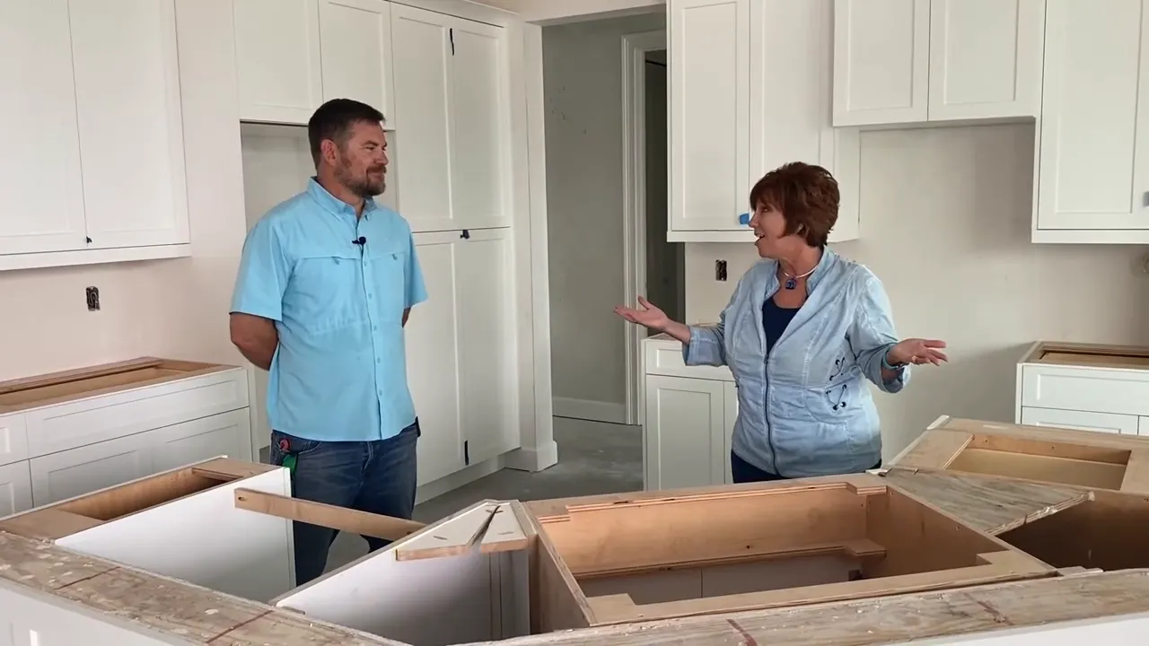 Wide mid‑construction kitchen shot showing white cabinetry, unfinished island boxes and two people discussing progress.
