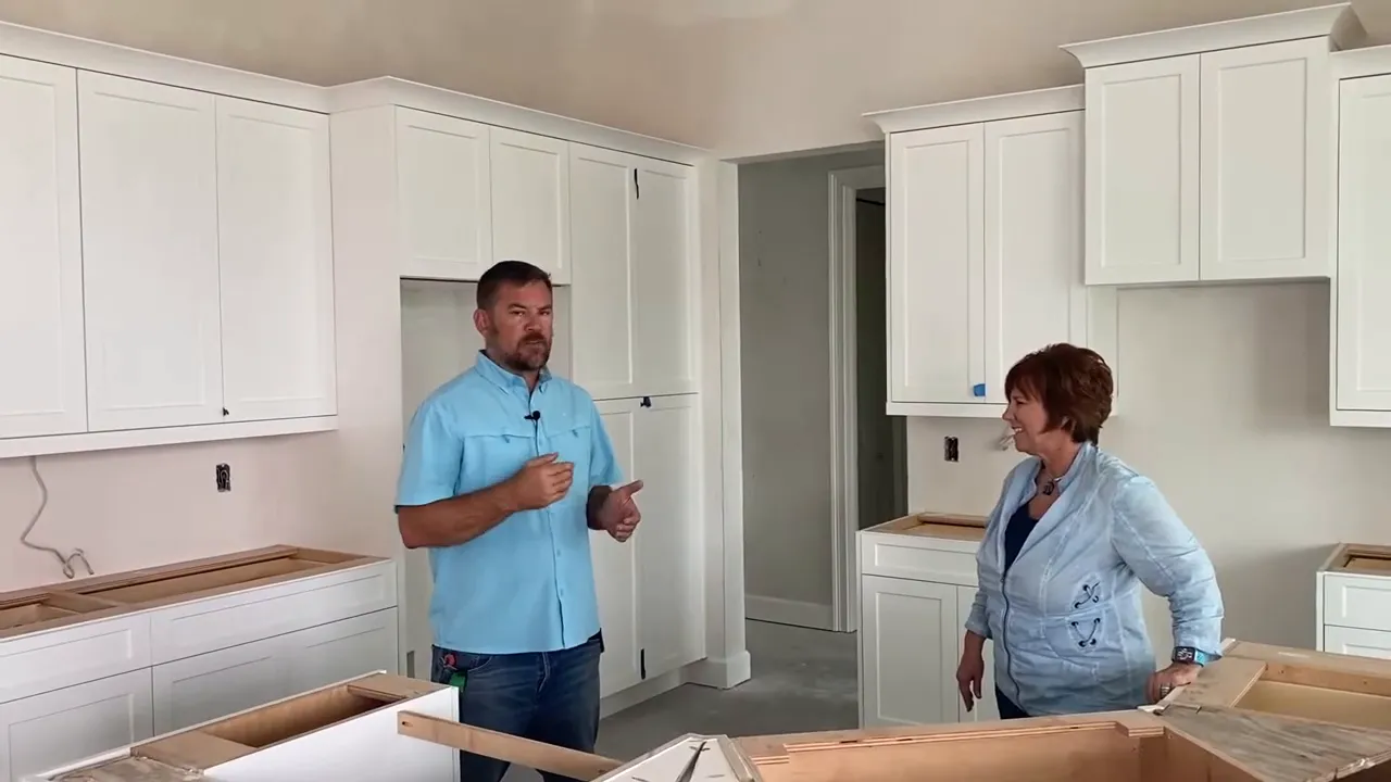 Mid-construction kitchen showing installed white cabinets, unfinished island boxes, and two people discussing progress.