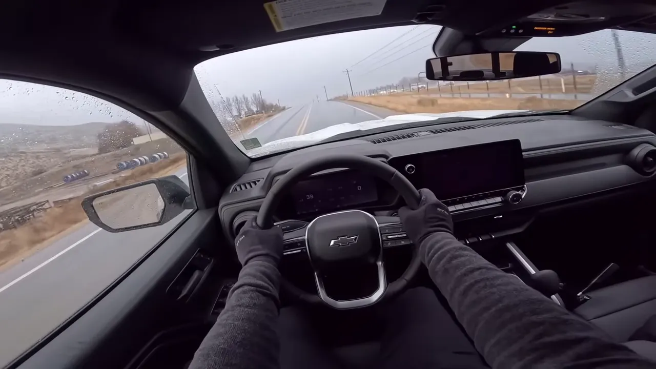 Driver's perspective inside a Chevrolet pickup showing the steering wheel with bowtie logo, dashboard and infotainment, with a clear wet road ahead and minimal wiper obstruction.