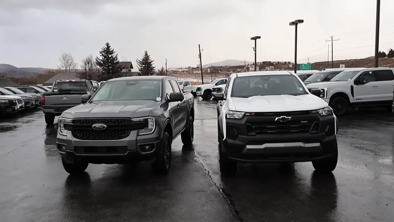 Front view of a gray Ford Ranger parked next to a white Chevrolet Colorado on wet pavement, showing both trucks' front-end styling and stance.