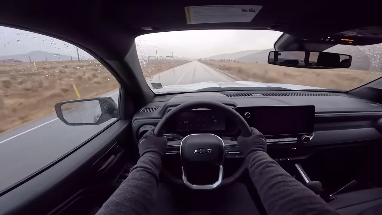 Driver's-seat perspective inside a Chevrolet pickup with an unobstructed windshield, light rain at the edges and a straight wet road ahead.