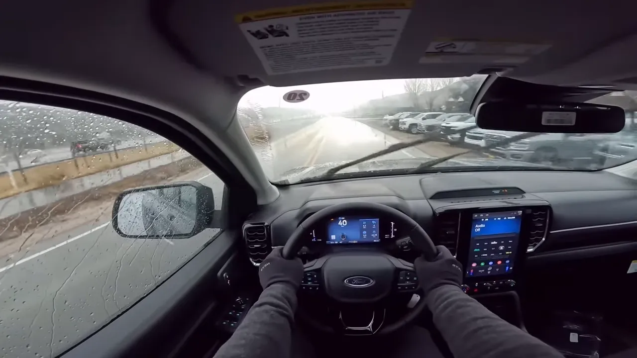 Clear point-of-view image from inside a Ford Ranger showing gloved hands on the wheel, the digital gauge cluster (40 mph) and the vertical infotainment screen with a rainy road ahead.