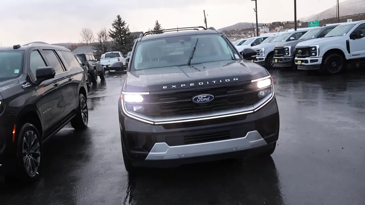 Front view of 2026 Ford Expedition Max showing grille, EXPEDITION hood lettering and illuminated headlights in a wet parking lot