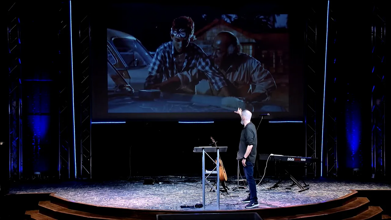 Speaker on stage pointing up at a projected Karate Kid scene while addressing the congregation