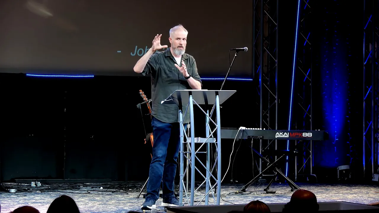 Pastor at lectern forming a rounded hand gesture as if holding something, clear stage lighting and keyboard visible