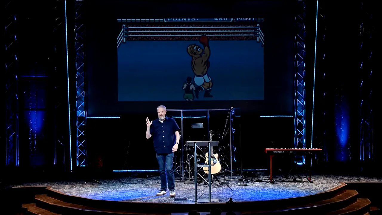 Speaker presenting on stage with a video-game graphic on a large screen during a sermon about discernment