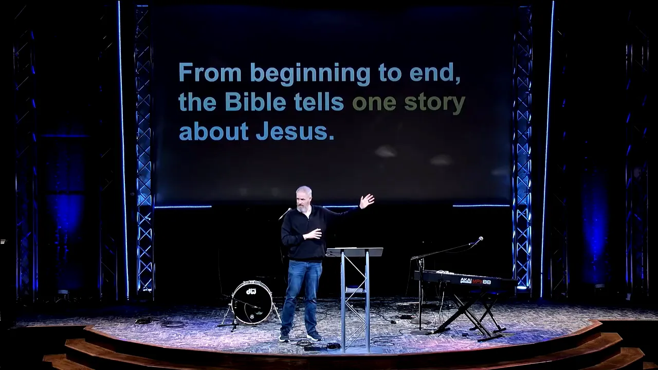 Wide shot of stage with slide text 'From beginning to end, the Bible tells one story about Jesus' and speaker gesturing.