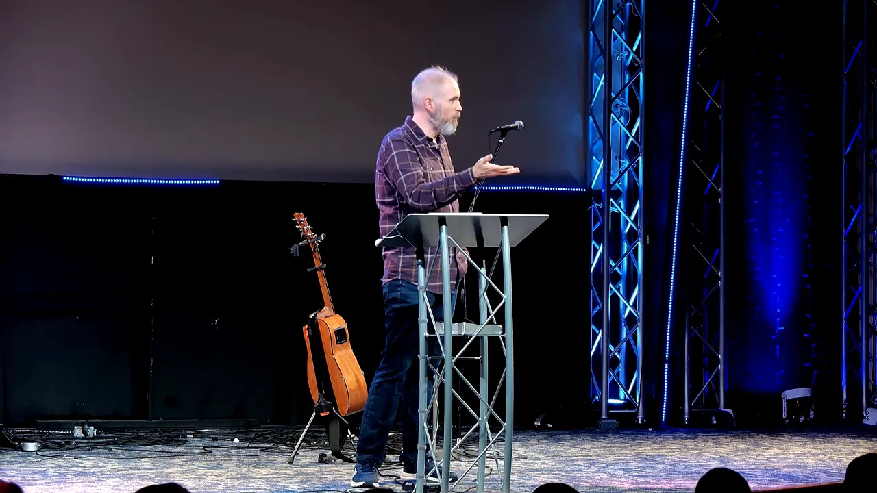 Speaker on stage at a lectern with an open palm gesture, guitar behind him and blue stage lighting.