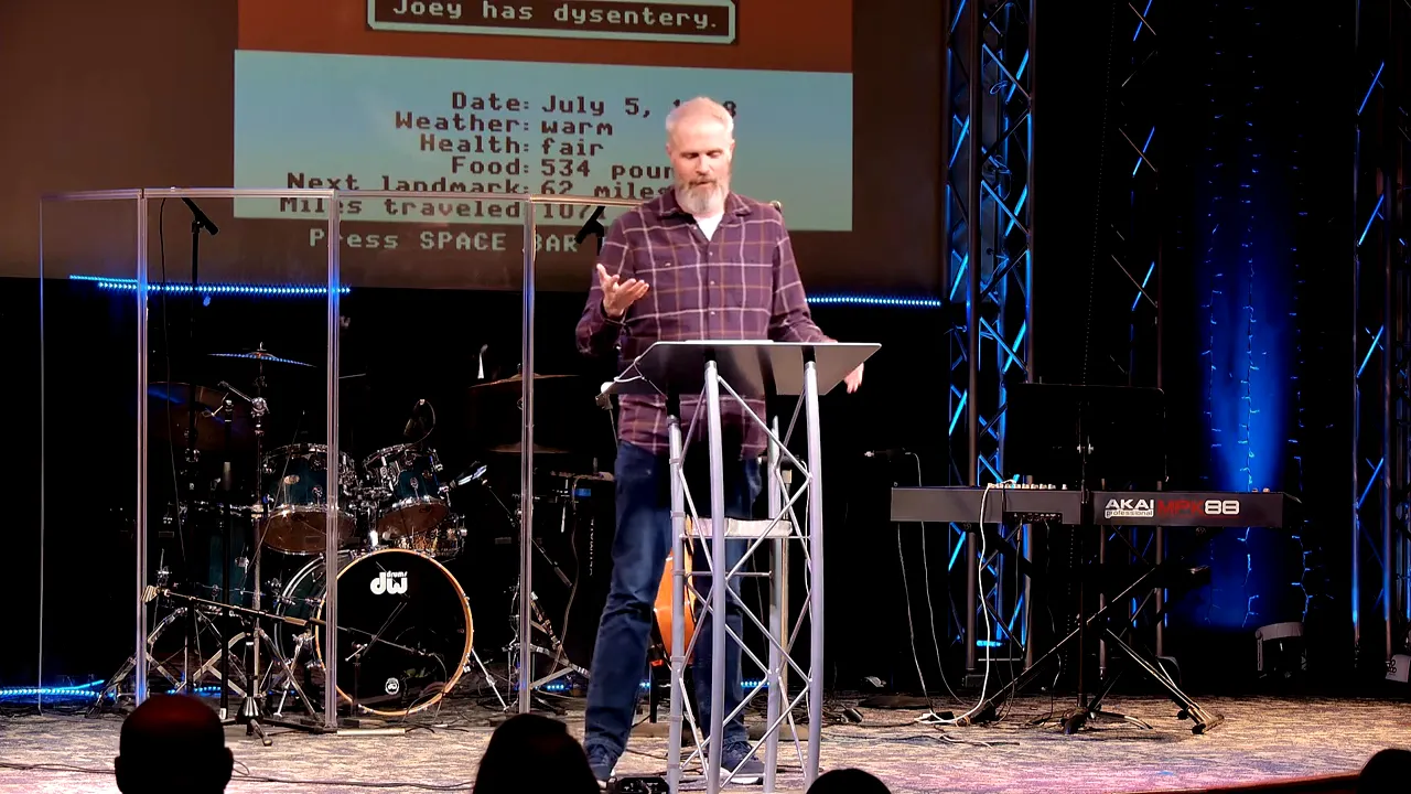 Speaker at a podium with an Oregon Trail game screen projected behind him showing game stats and the message 'Joey has dysentery'.