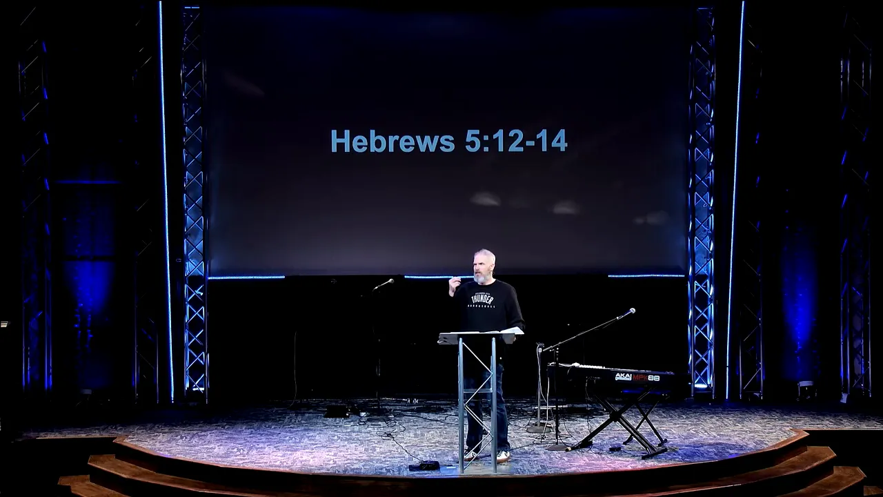 Wide stage shot of the pastor gesturing at the audience with a large 'Hebrews 5:12-14' slide overhead.