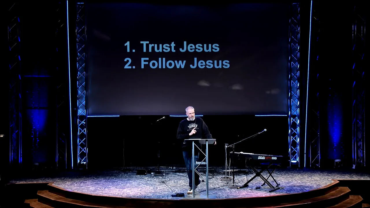 Stage slide that reads '1. Trust Jesus 2. Follow Jesus' with a speaker at a podium