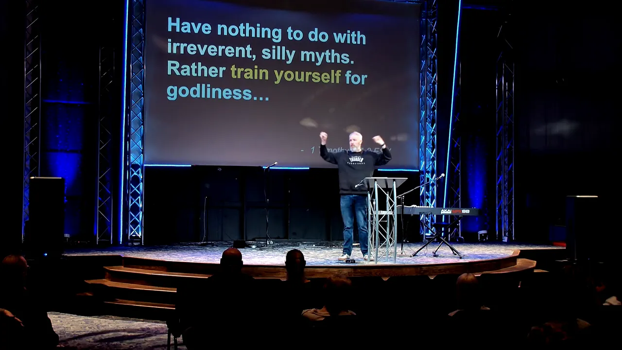 Stage with projected text 'Rather train yourself for godliness' and speaker raising both hands at the podium.