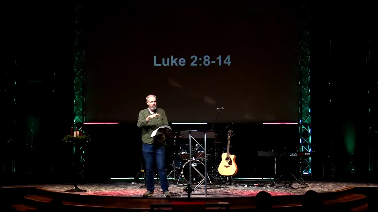 Speaker on stage with 'Luke 2:8-14' projected on a screen, guitar and drum kit visible in background