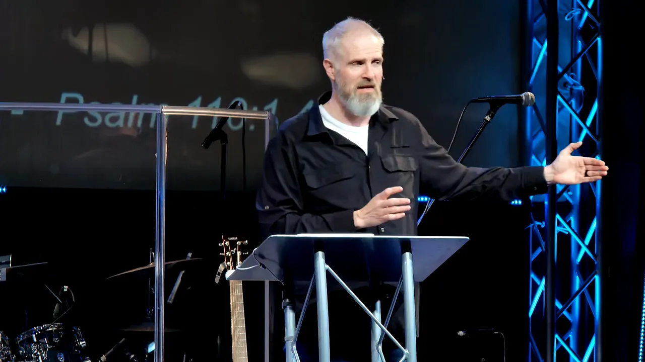 Speaker beside a lectern with his arm extended and the words 'Psalm 110:1,4' visible on the screen behind him