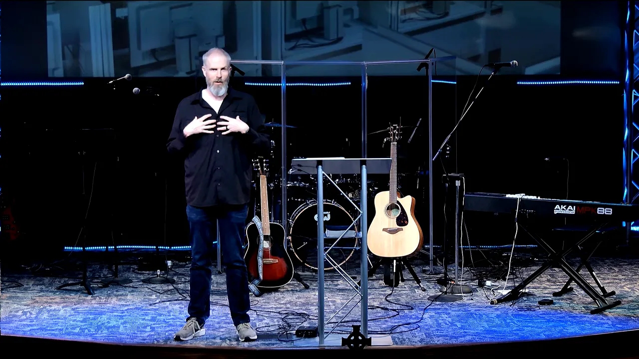 Full-stage shot with speaker centered behind the podium area, guitars and keyboard visible on stage