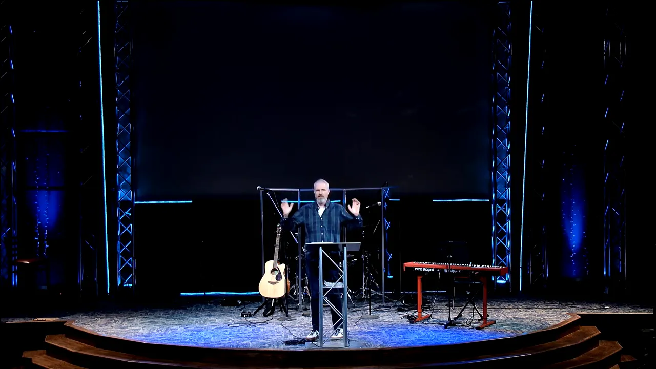 Pastor standing at a podium with both hands raised on a church stage with blue lighting and instruments