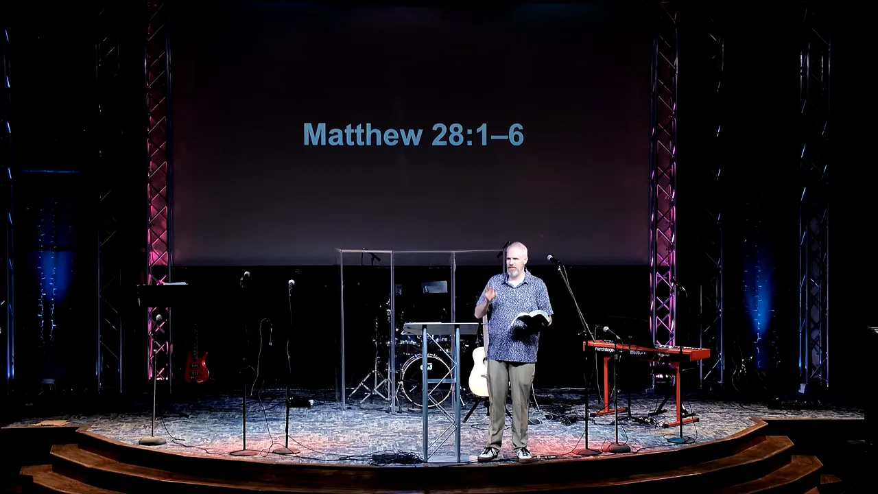 Preacher speaking with hand raised while holding a Bible on stage with Matthew 28:1–6 displayed