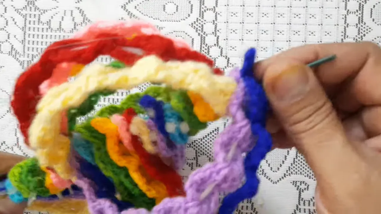 Hands gathering blue and lavender strip ends onto a metal support beside a rounded multicolored crochet structure