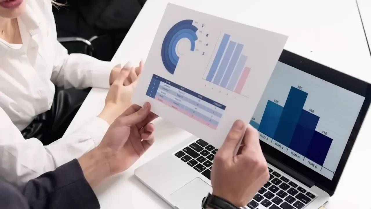 Two people reviewing printed charts while a laptop displays a bar chart.