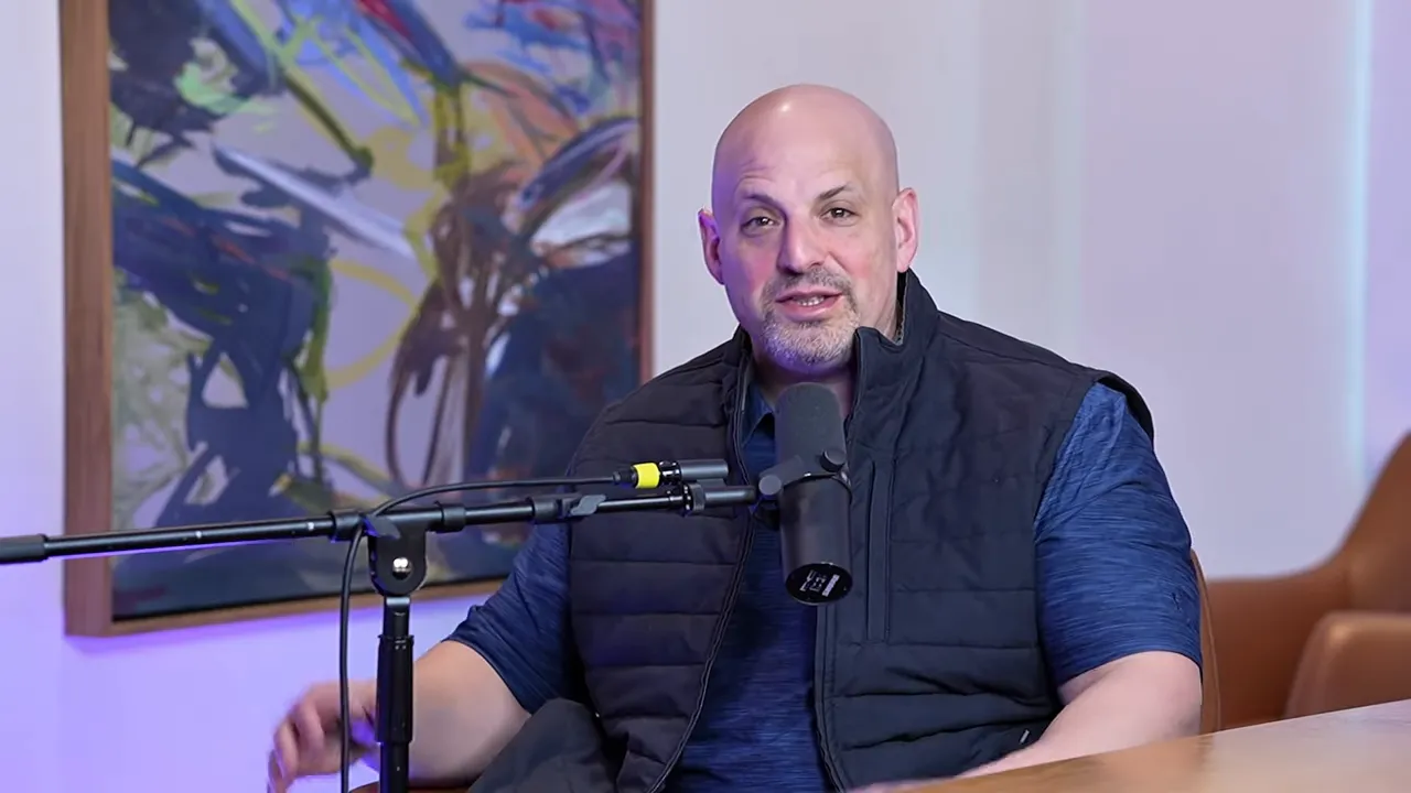 Clear mid-shot of podcast host seated at a table speaking into a microphone with studio artwork behind him