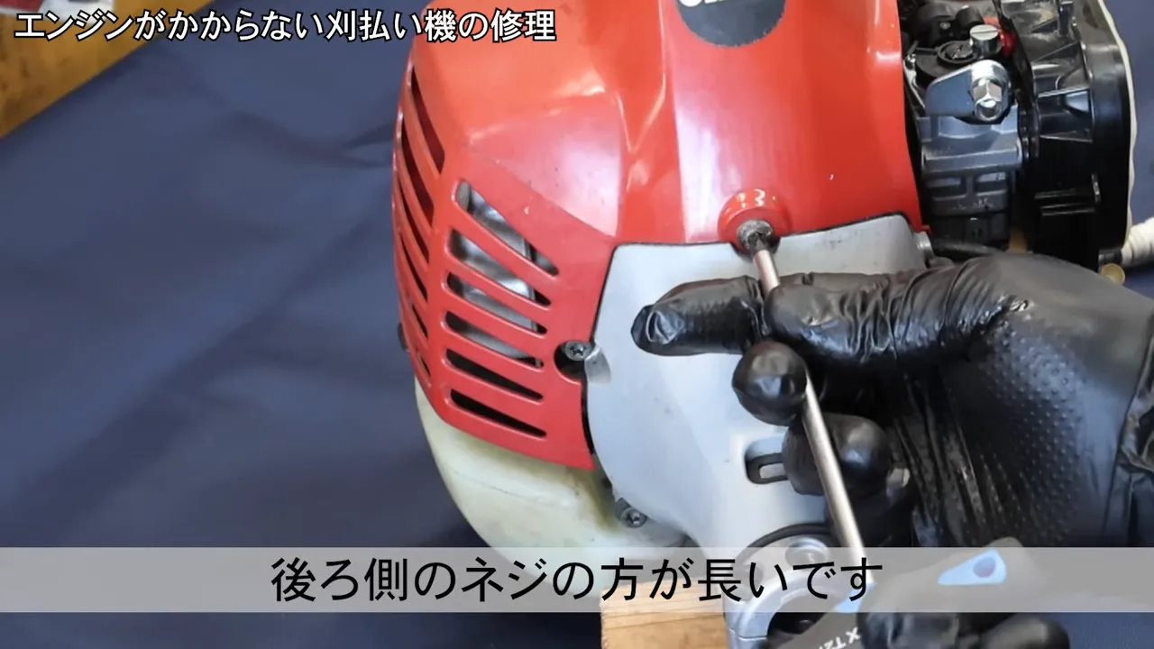 黒い手袋でドライバーを使い、エンジンカバー後側のネジを外す場面（後ろ側のネジが長いことを示す）