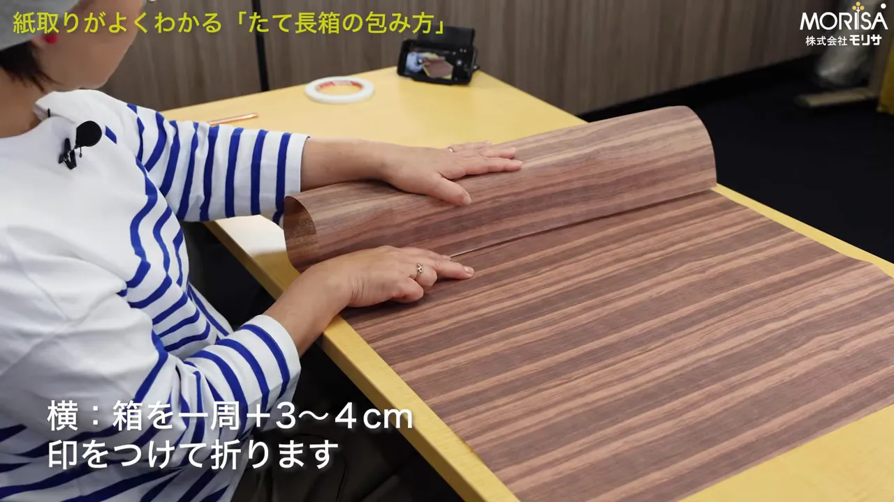 包装紙の上で指先が折り目の位置を示している。画面下に「横：箱を一周＋3〜4cm 印をつけて折ります」のテロップ