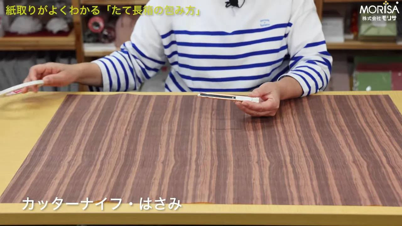 木目の包装紙の上でカッターナイフを手に持つ準備をする様子（カッターナイフ・はさみの表示あり）