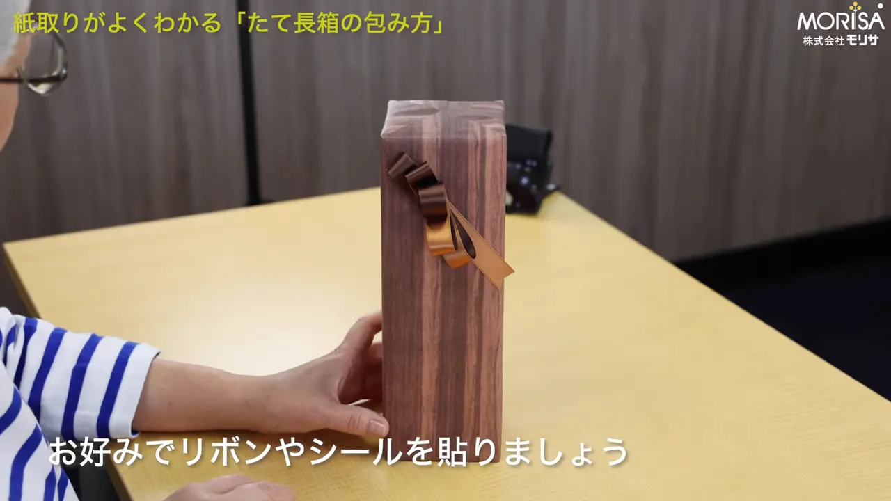 木目のたて長箱にブラウンとゴールドのリボンを飾った完成イメージ。画面下に「お好みでリボンやシールを貼りましょう」のテロップ。