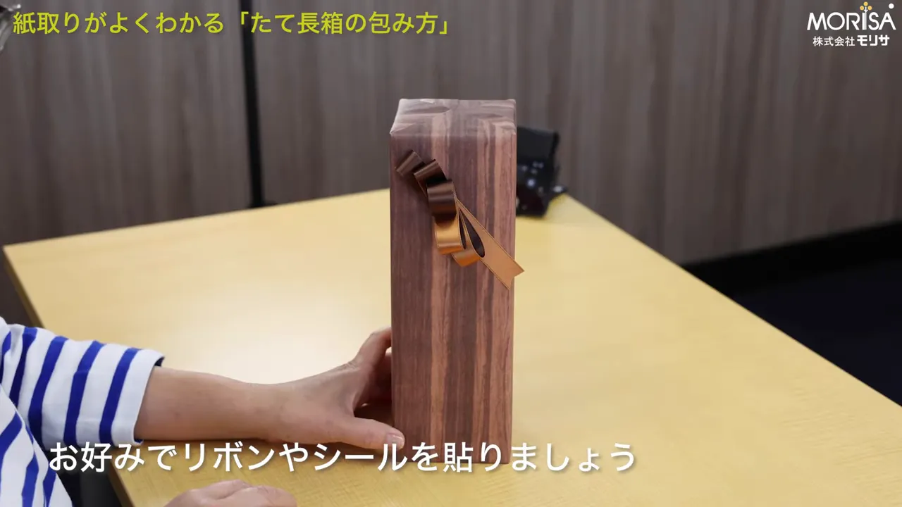 木目の包装紙で包まれたたて長の箱の正面がはっきり見え、上部に波形リボンが付いた完成品。手が箱の下部を支えている。