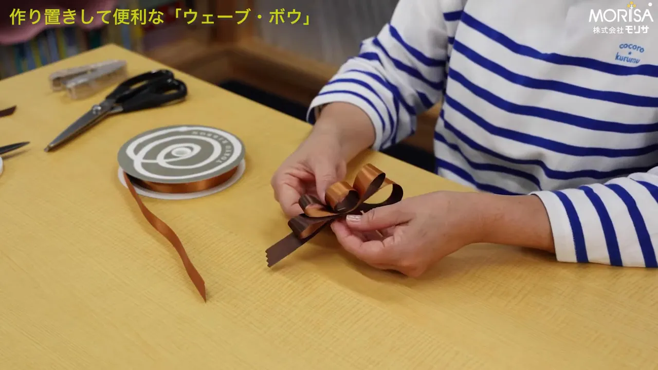 ホチキスとリボンスプールが見えるテーブルでウェーブ・ボウの中心を押さえている手元