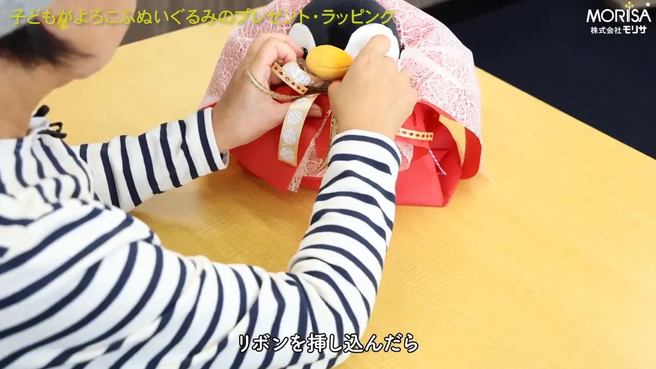 リボンを差し込みぬいぐるみの仕上げをする手元のクローズアップ（字幕あり）