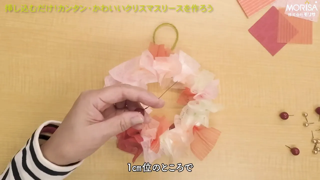 上から見た作業風景。小さな発泡スチロールリースに赤いベリーピックを差し込もうとする手元と、右側に並べられた予備のピックが見える。