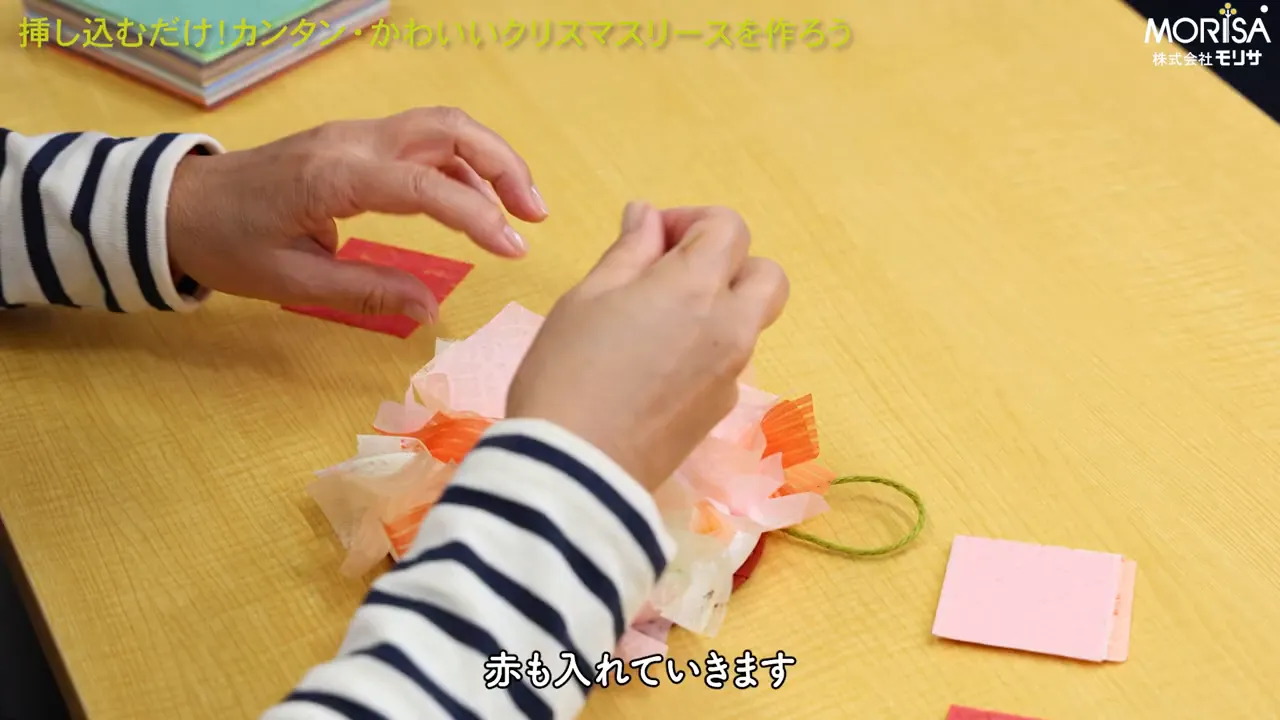 ボーダーの袖の手が色とりどりの和紙の小片を発泡スチロール製リースに挿し込んでいる上からの手元写真