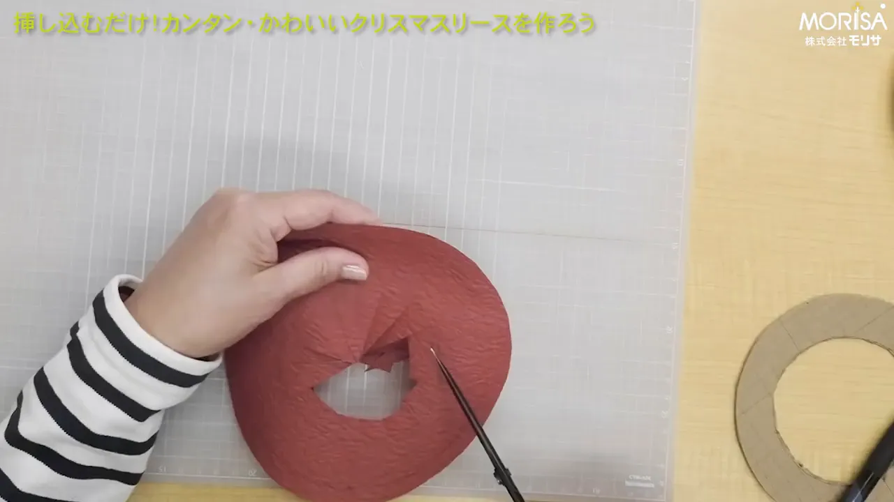 上から見た作業台で赤い和紙を発泡スチロールリースに合わせ、手で折り返して整えている手元のクローズアップ。段ボールの土台とカッターマットが見える。